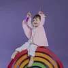 Enfant déguisé en licorne rose, assis sur un arc-en-ciel en bois coloré, tenant une bouteille sensorielle violette.