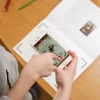 Enfant tenant un smartphone scannant un cahier dessin animé Léonard De Vinci sur bureau en bois.