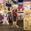 Deux figurines en carton debout, femme aux cheveux blancs en tailleur marron, enfant blonde avec pancarte TBDIB, décor de tra