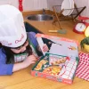 Enfant avec toque blanche Chefclub et tablier à carreaux devant coffret Kids Le Pizzaiolo ouvert.