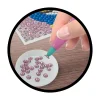 Main tenant un stylet violet et vert, posant un strass rose sur toile Glitters Paon Buki.