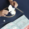 Mains assemblant un papillon blanc en feutrine avec décorations bleues et instructions papier.