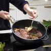 Poêle noire Cookut 28 cm avec steaks, ail et branche de romarin en cuisson.