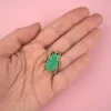 Pin's scarabée vert pailleté avec contour doré tenu dans une main sur fond rose.